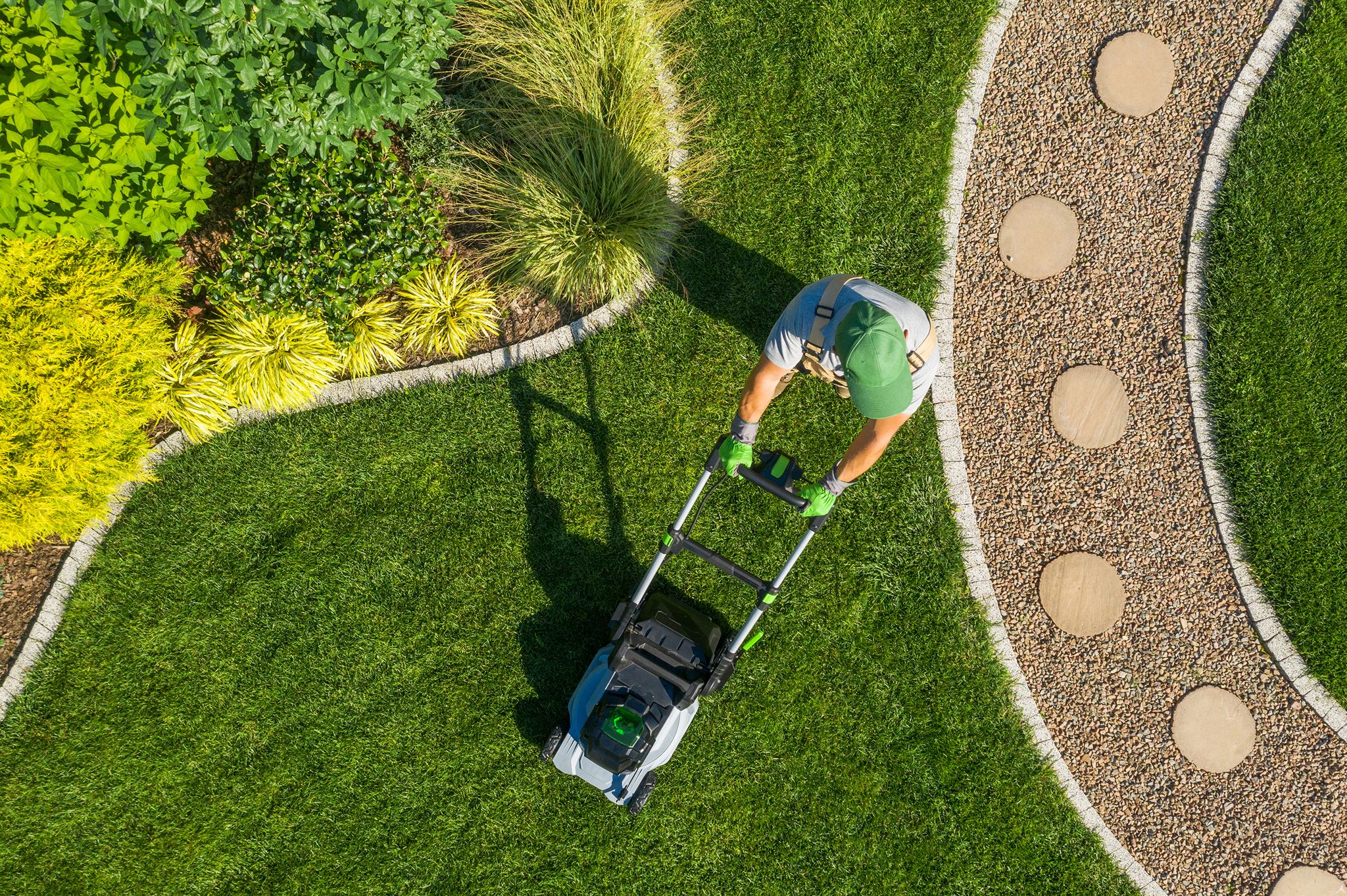 Aerial-view-of-person-pushing-lawn-mower-on-grass-between-garden-bed-and-path